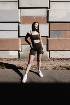 Confident woman in fashionable athletic wear posing against an industrial wall under sunlight.
