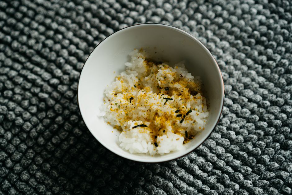 A small jar of furikake seasoning tipped over a bowl of steamed white rice, showing flecks of dried seaweed, sesame seeds, and pink and white flakes scattered across the grains