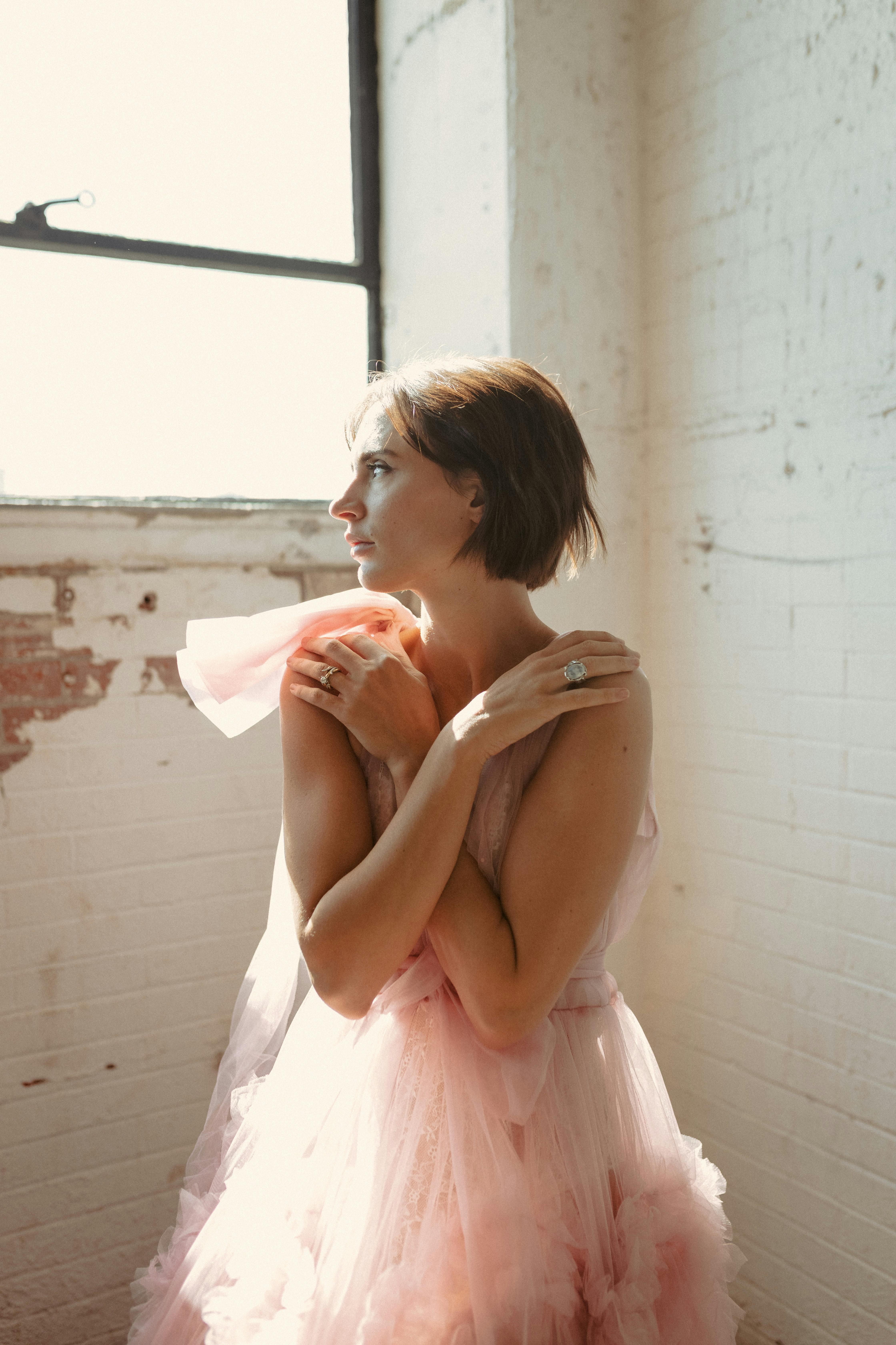 A fashionable woman in a pink dress poses gracefully in a sunlit brick room.