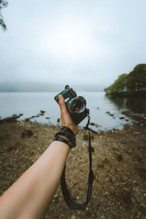 Free Person holding a camera near a serene lake in Cumbria, England, capturing the scenic landscape. Stock Photo