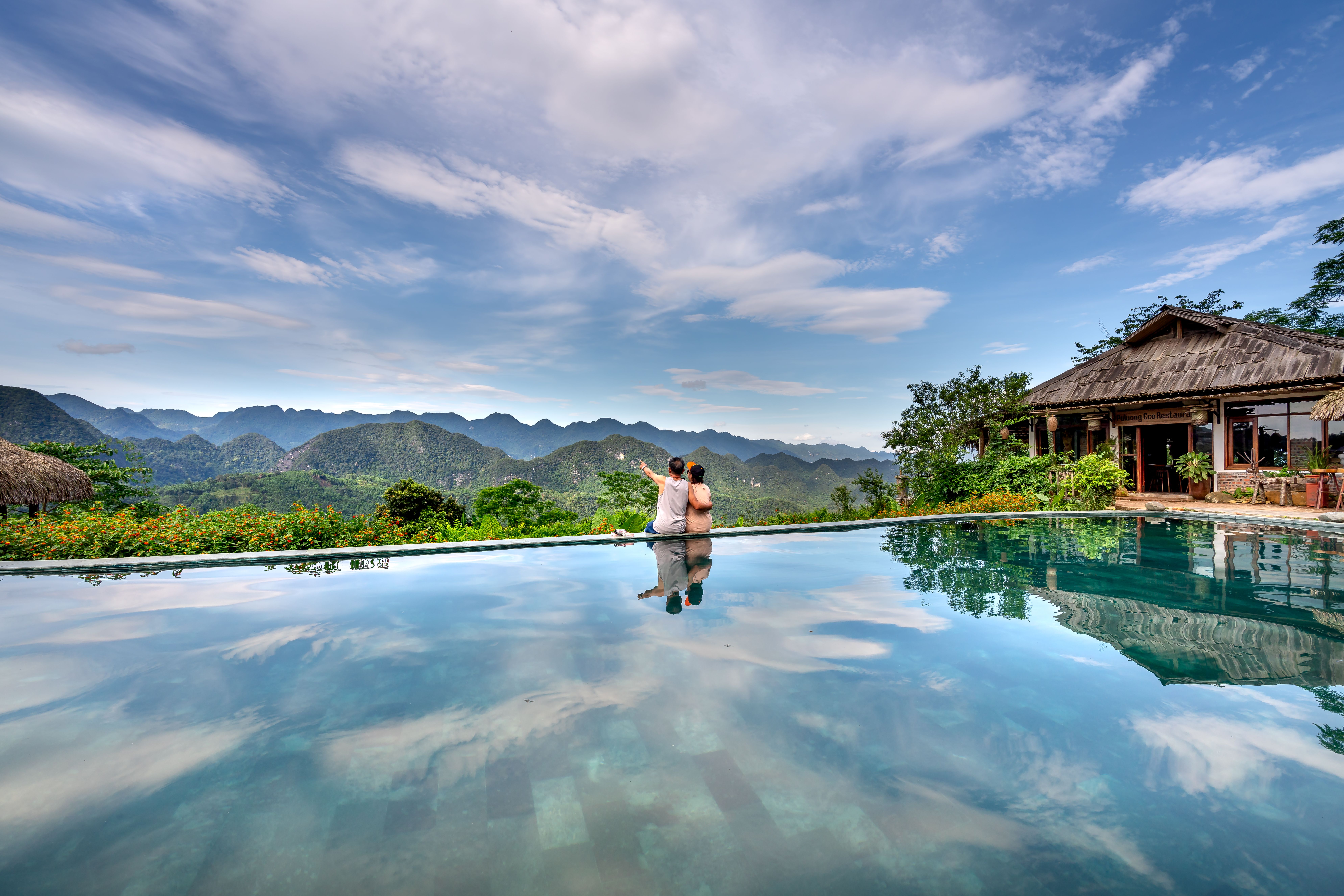 Casal desfrutando de vista panorâmica de uma piscina infinita, com montanhas e céu azul, em refúgio de luxo.