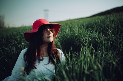 Free A woman sits in a lush green field wearing a striking red hat, embracing nature's beauty. Stock Photo