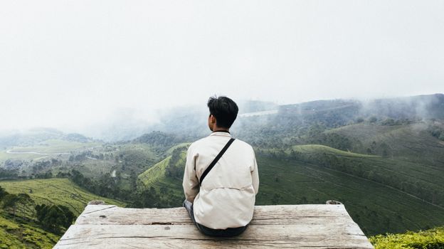 Man sitting on a wooden deck, enjoying the serene view of lush green valleys under misty skies.