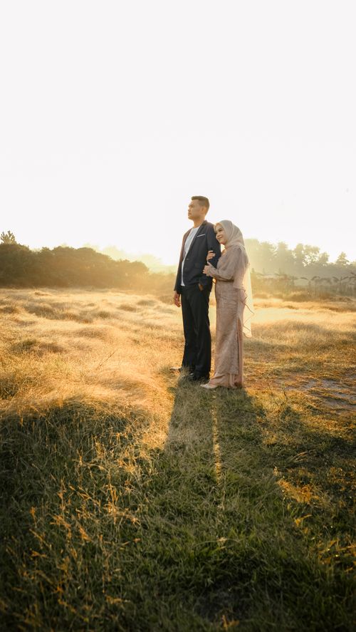 Free A couple stands together in a grassy field at sunset, embracing with love and warmth. Stock Photo