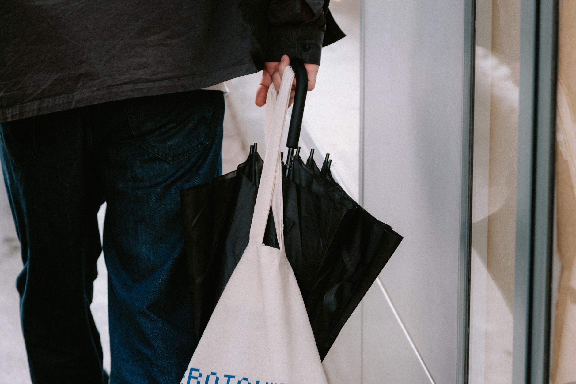 Man holding a black umbrella and canvas tote bag, walking on a city sidewalk.