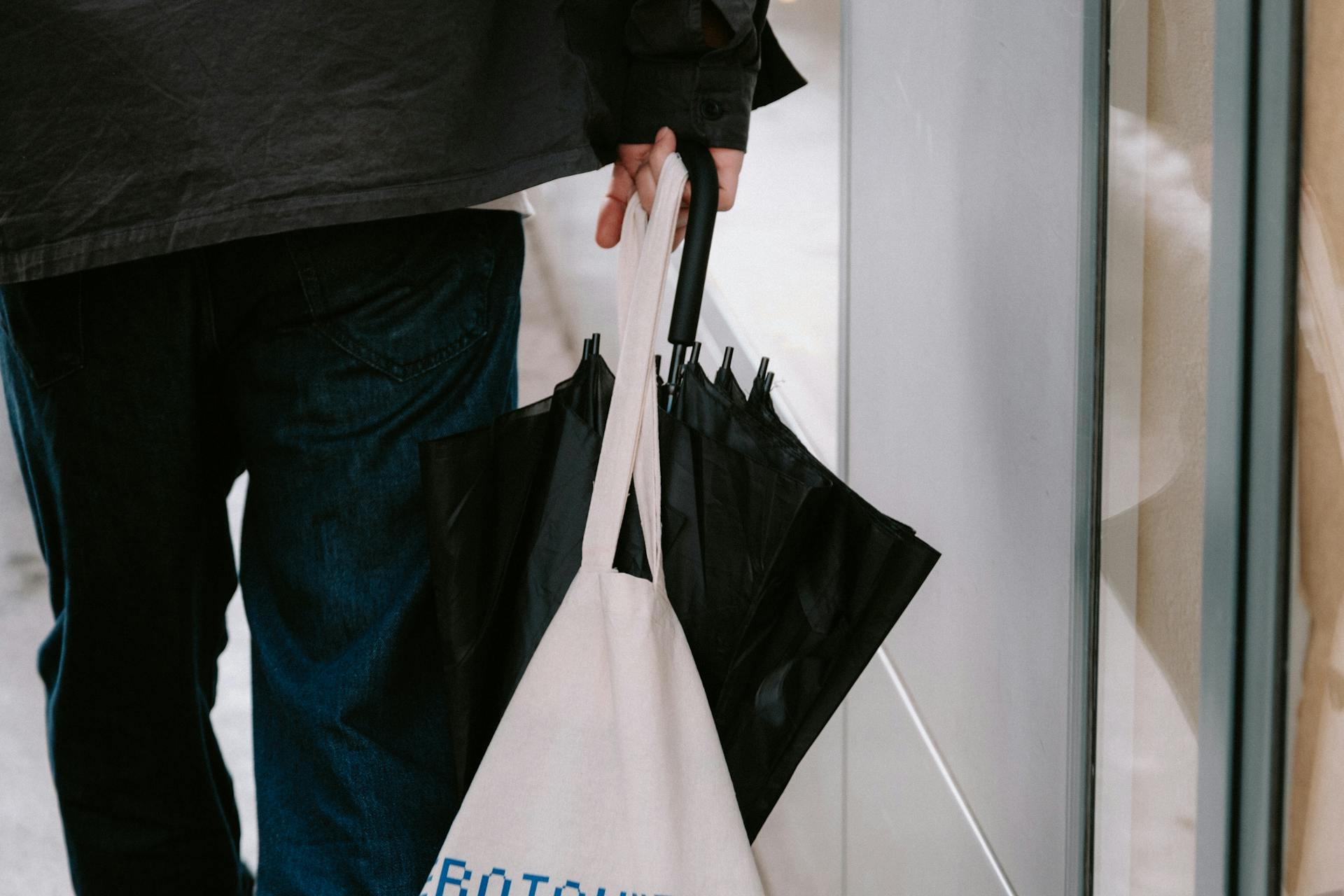 Man holding a black umbrella and canvas tote bag, walking on a city sidewalk.