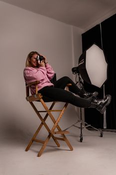 Young woman in pink hoodie taking photos in a studio setting, relaxed on a director's chair.
