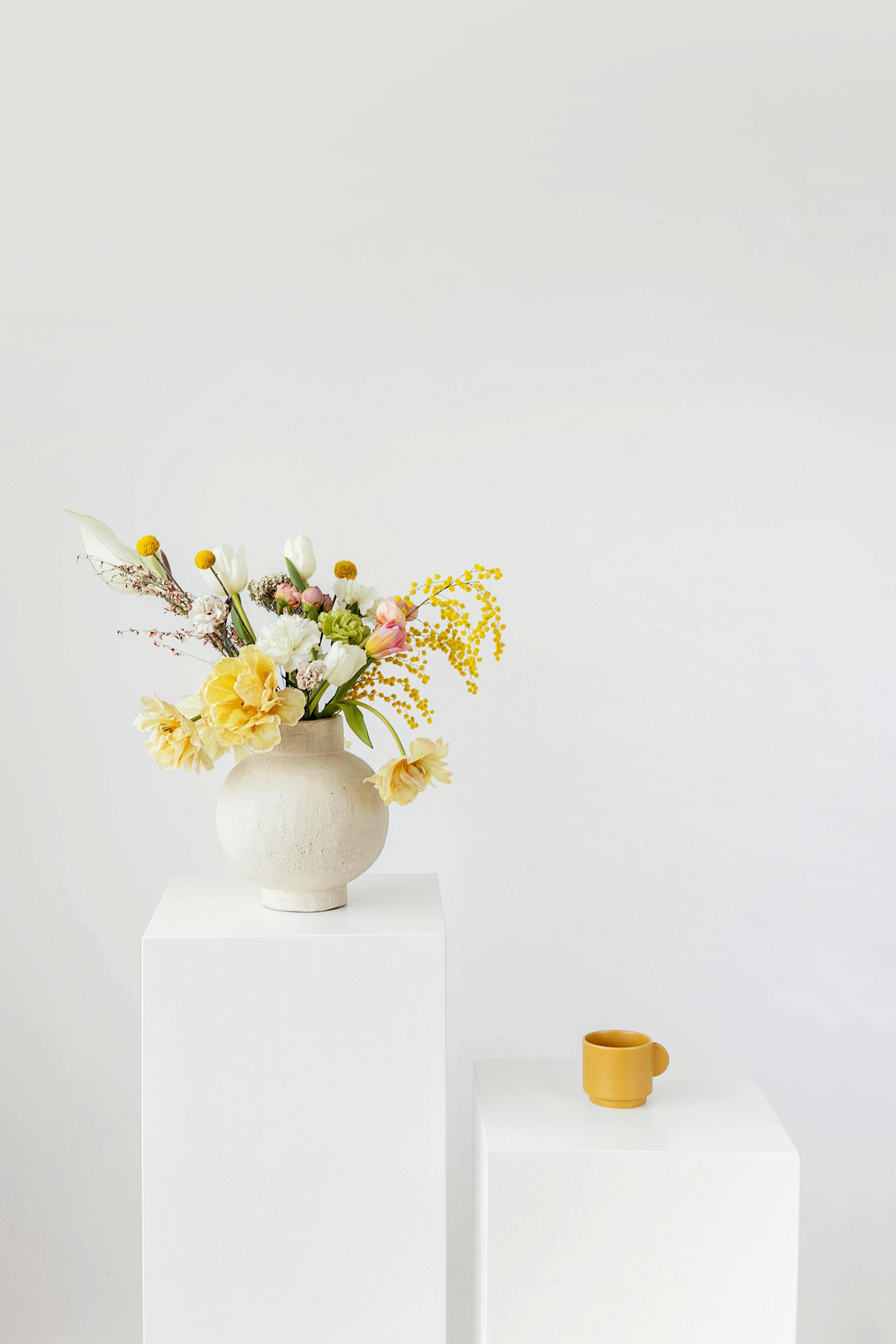 A minimalist floral arrangement in a vase beside a mug on white platforms with a white background.