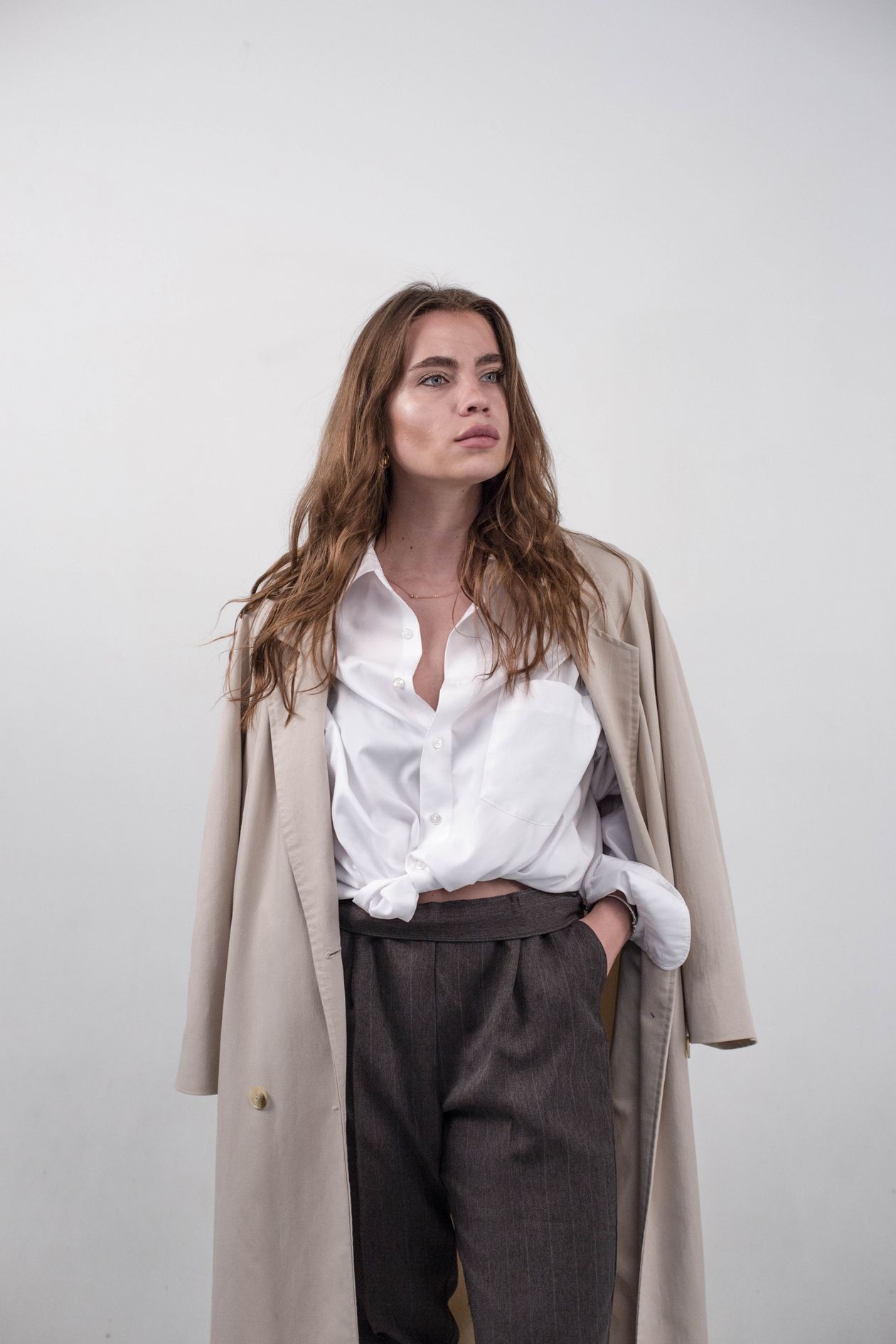 Woman in a white button-up shirt, beige trench coat, and gray trousers.