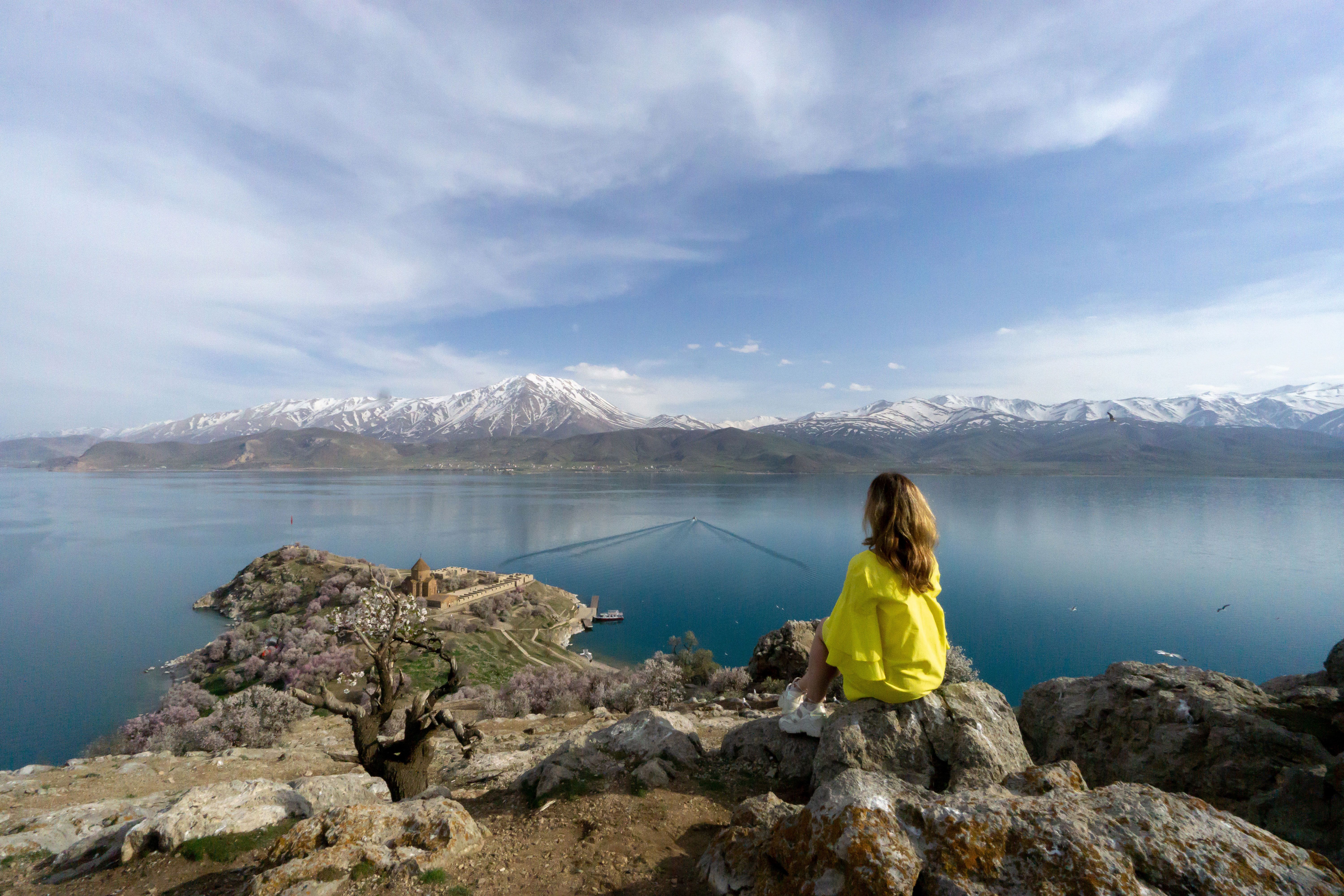 Woman in yellow overlooks Akdamar Island’s stone church, Lake Van and snowy mountains in Van, Turkey, under soft daylight