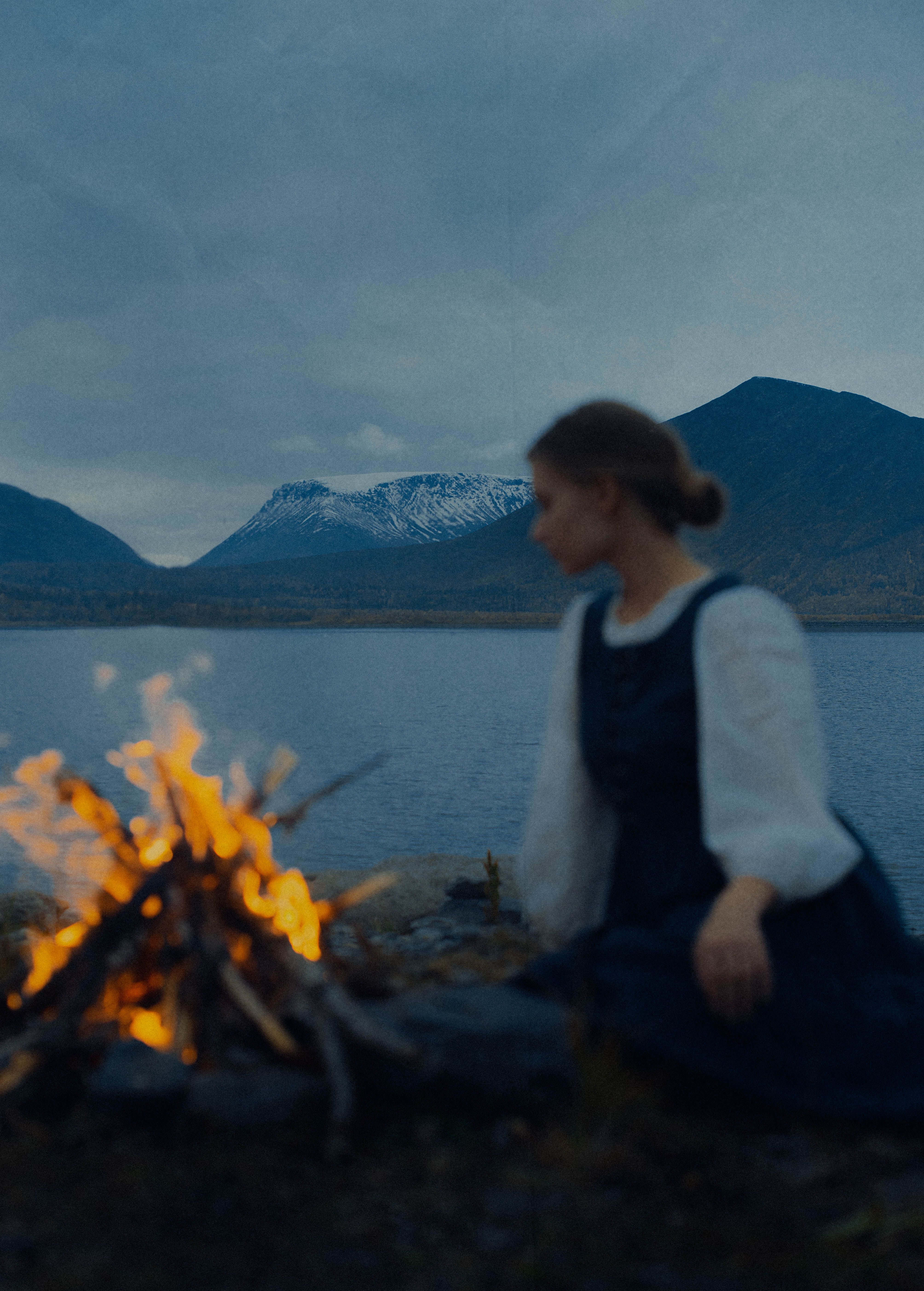 A woman sitting by a bonfire near a serene mountain lake at twilight.