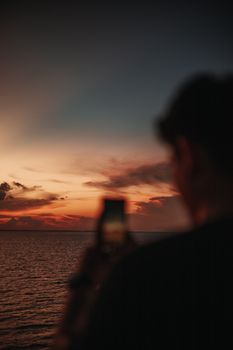 Silhouette of a man photographing a dramatic sunset over the ocean with a smartphone.