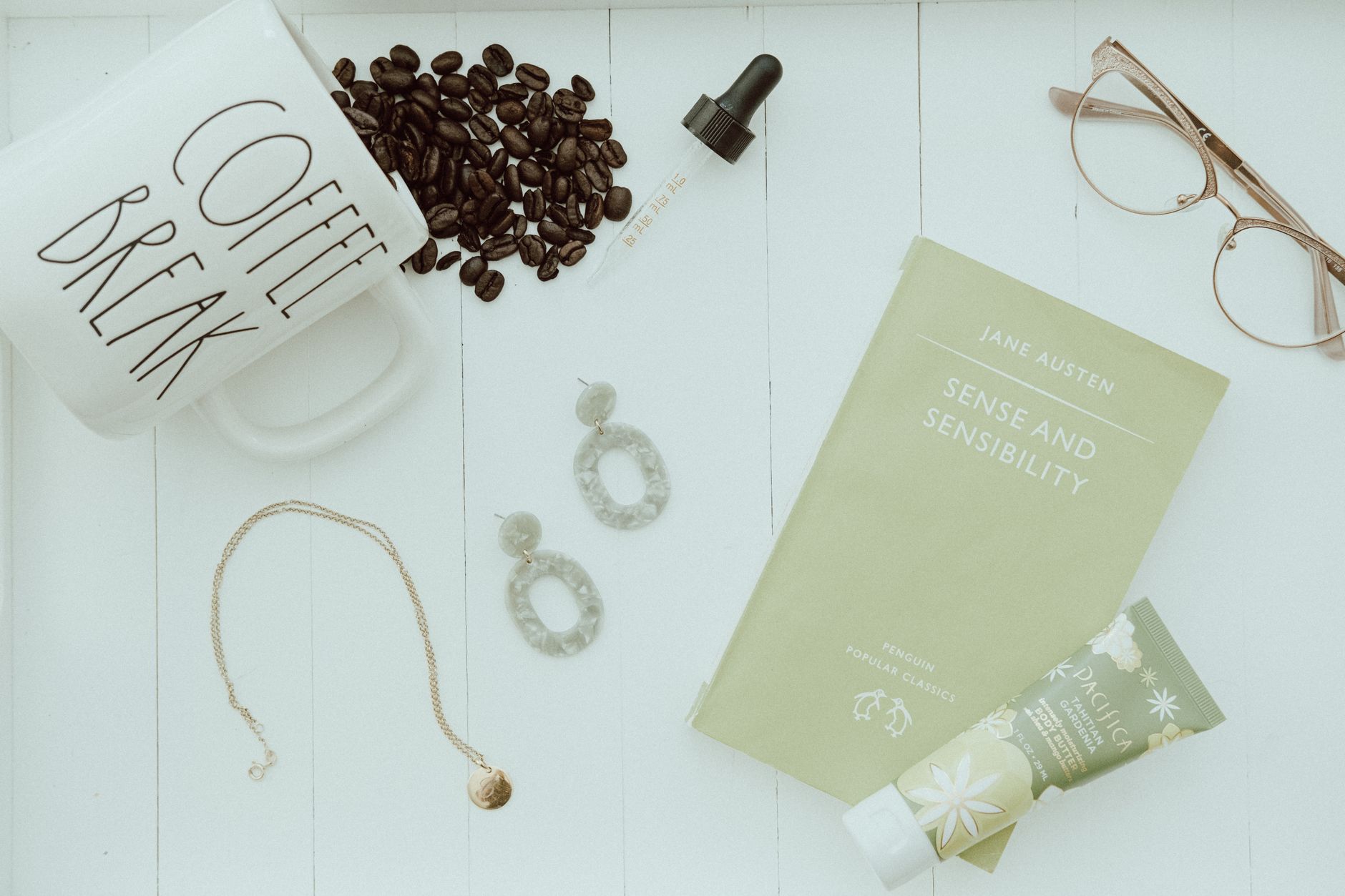 A minimalist flat lay featuring coffee, a book, jewelry, and eyeglasses on a white surface.