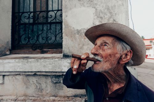 Free Elderly man enjoying a cigar in the street, wearing a straw hat and showcasing character with wrinkles. Stock Photo