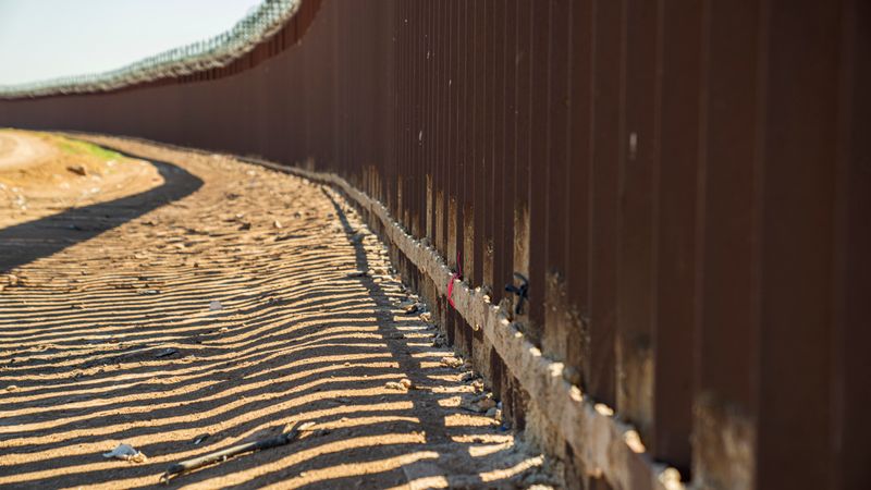 A long metal border fence casting shadow patterns on a sandy desert path in bright sunlight.