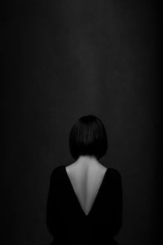 A dramatic, minimalist portrait of a woman from behind in a black dress with open back against a black background.