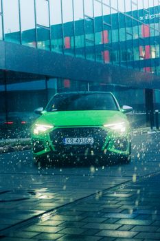 A vibrant green luxury car with headlights on, parked in a rainy urban environment.