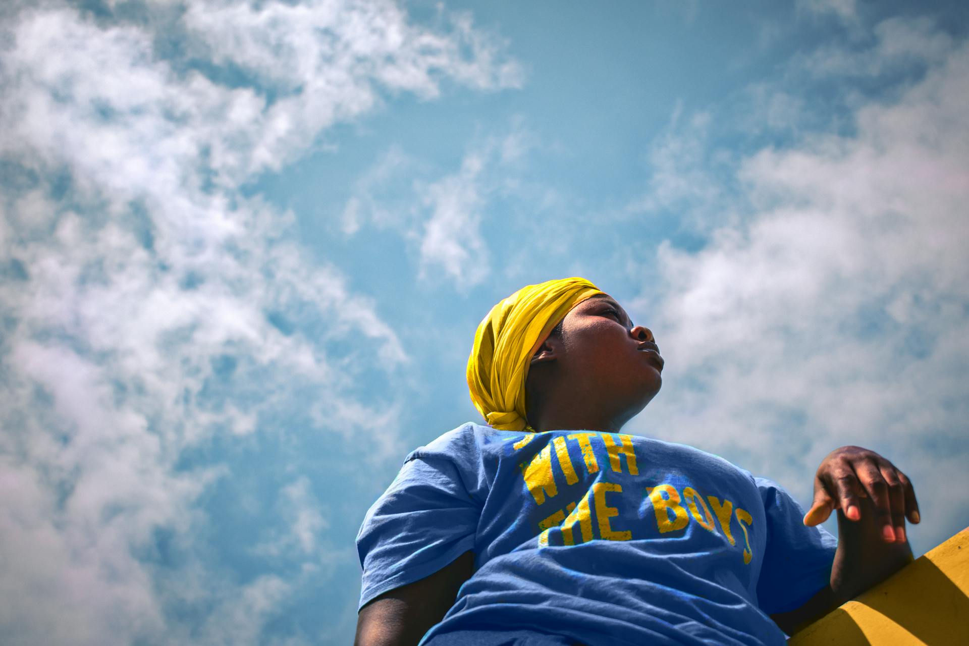 A young woman in a blue shirt and yellow headscarf gazes upwards against a bright Ghanaian sky.