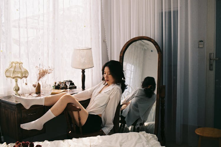 Brunette Woman In Bedroom Sitting In Chair with Legs On Backrest