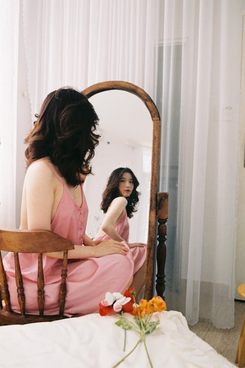 A Woman in Pink Dress Sitting on a Wooden Chair while Looking at the Mirror