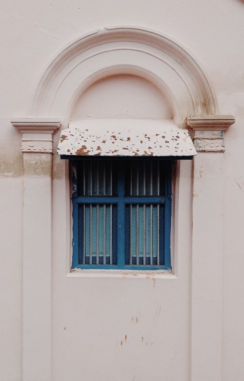 Free A classic blue window with shutters set in a beige wall, featuring an ornate arched design and peeling paint. Stock Photo