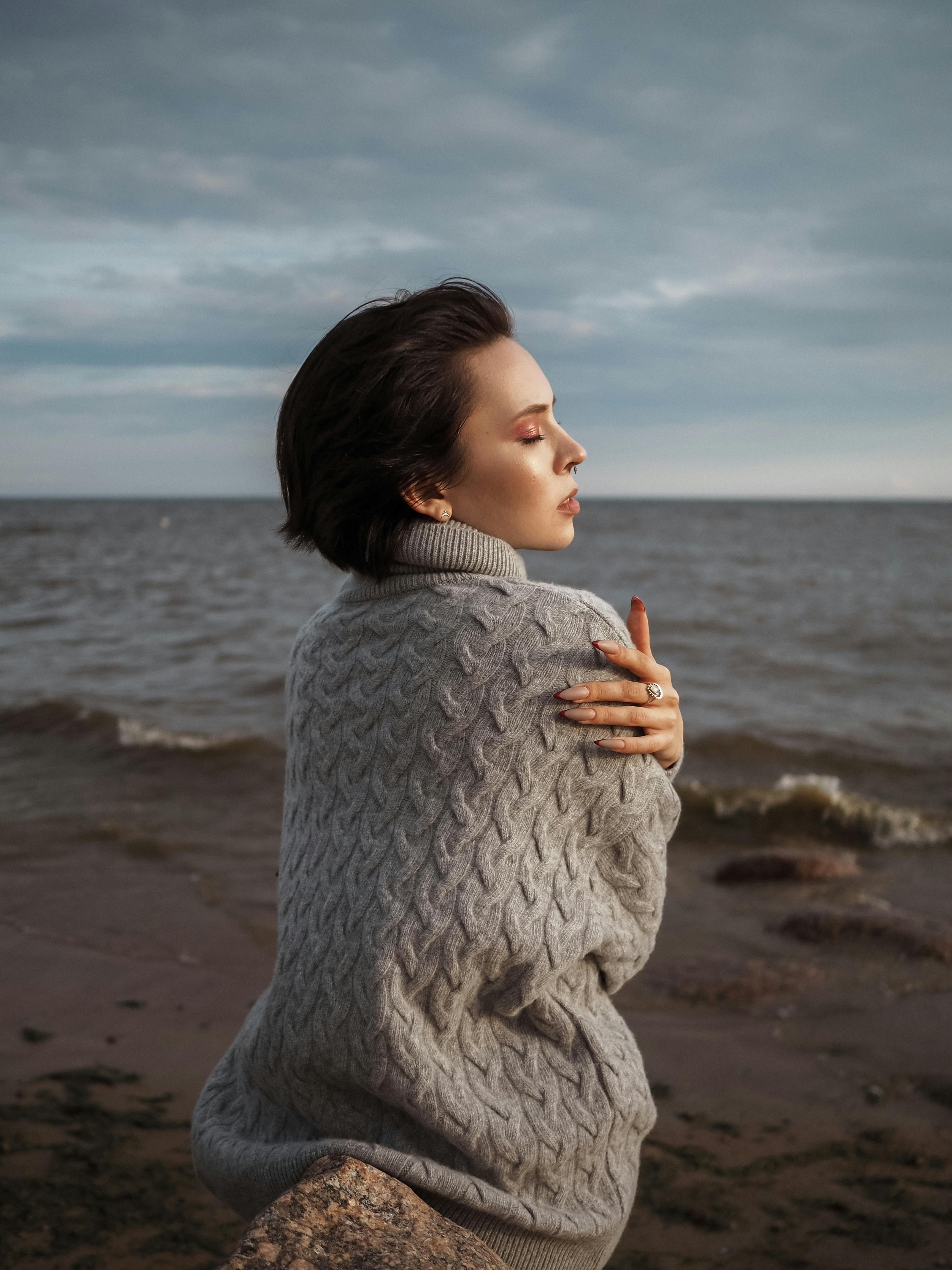 一位沉思的女人穿着舒适的毛衣，坐在海边，拥抱着平静的海风
