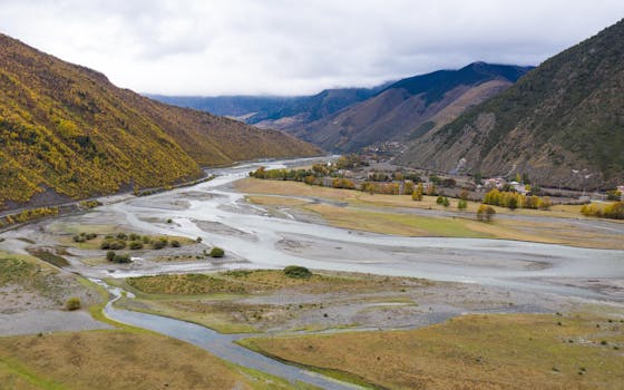 A panoramic view of a lush valley with a winding river and mountains under a cloudy sky.