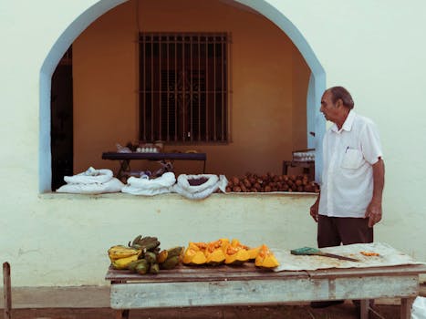 Senior man in white polo at outdoor market stall with assorted fruits and vegetables.
