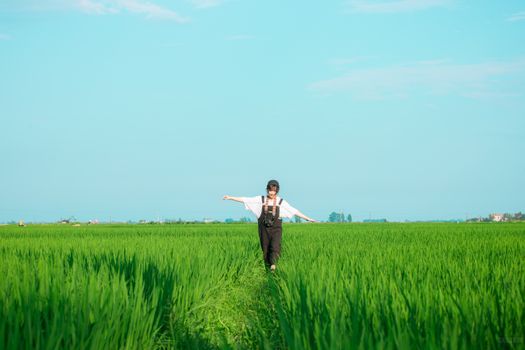 A young woman walks with arms outstretched in a lush, sunlit green rice field under a clear blue sky.