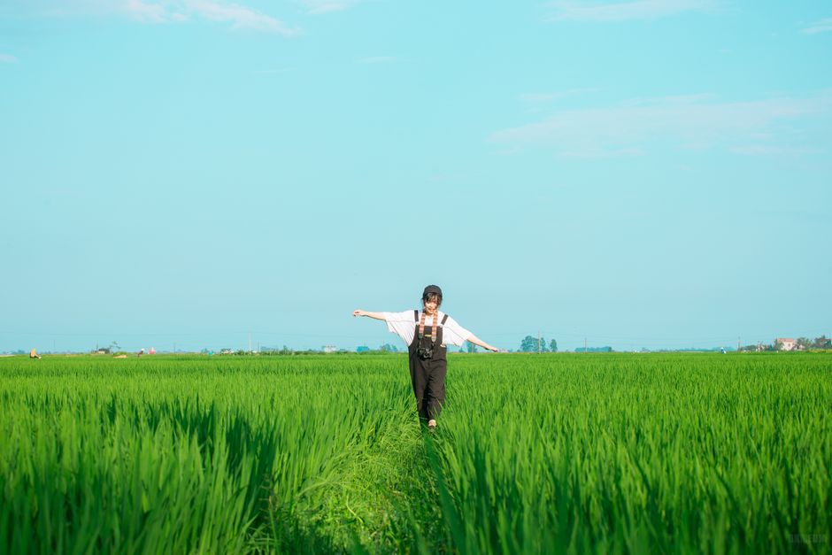 A young woman walks with arms outstretched in a lush, sunlit green rice field under a clear blue sky.
