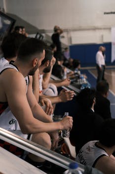 Basketball players in jerseys sitting on the bench during a game at an indoor gym.
