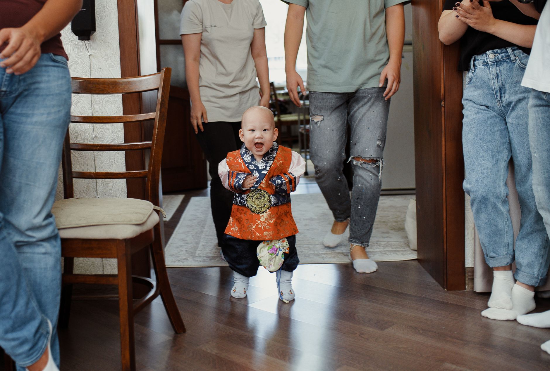 A joyful baby in traditional attire takes first steps with family support in a cozy indoor setting.