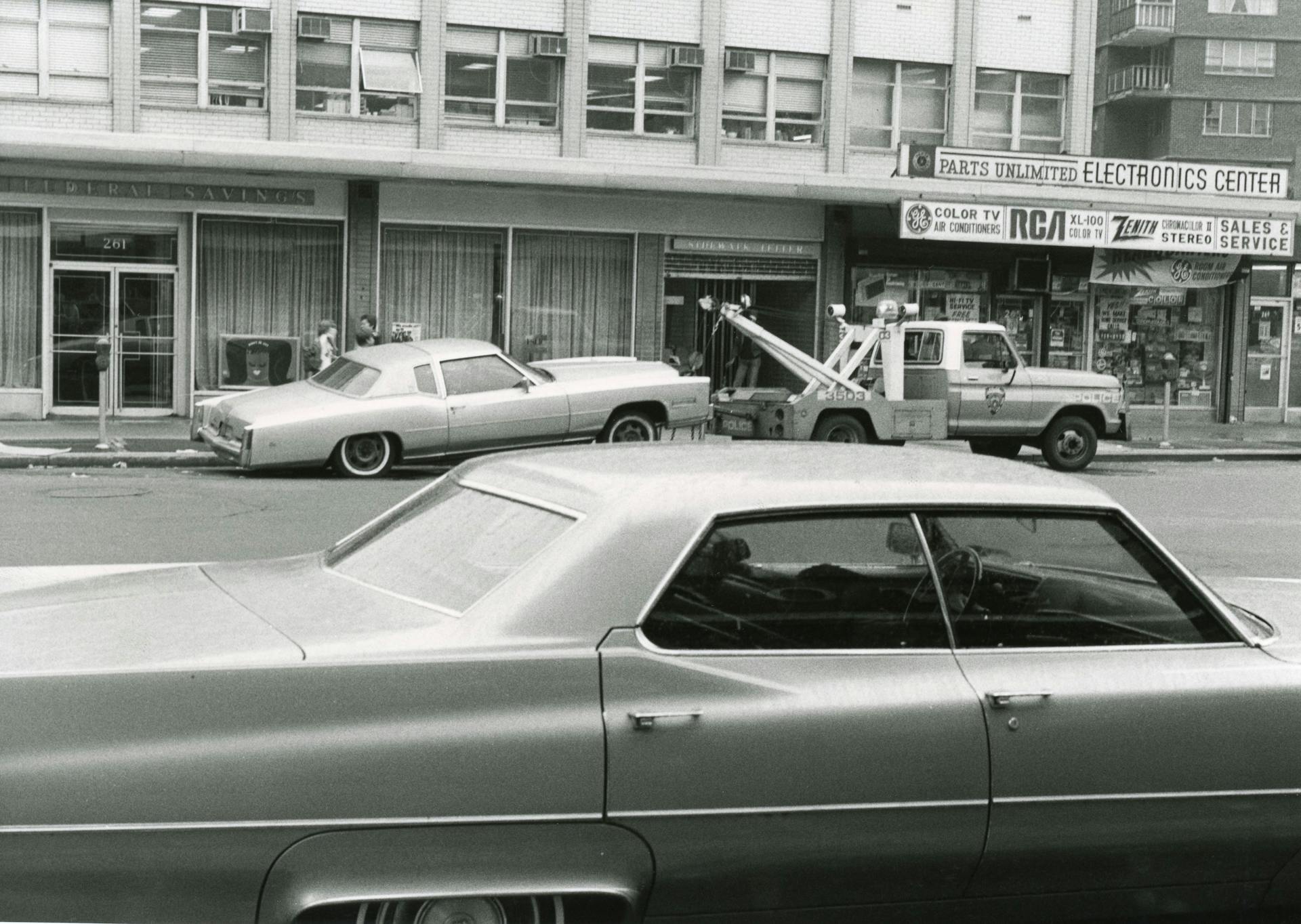 Black and white photo of vintage cars with a tow truck in an urban street setting.