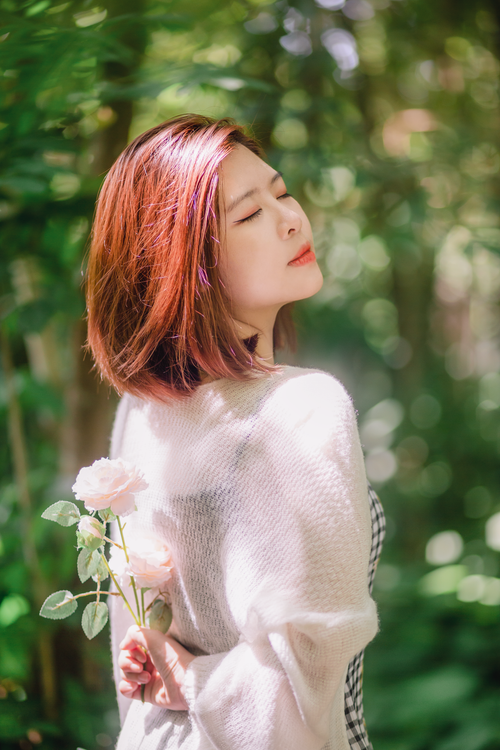 Free A woman enjoys a peaceful moment with eyes closed, holding a rose in a sunlit garden. Stock Photo