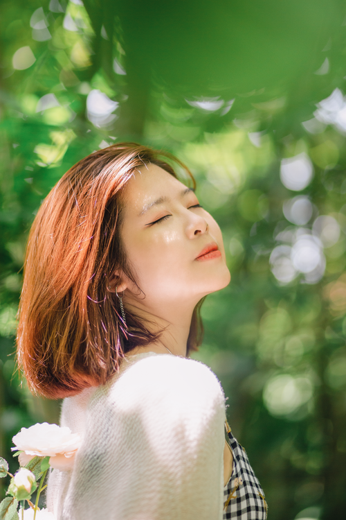Free A woman enjoys the warm sunlight in a lush garden, eyes closed, holding a rose. Stock Photo