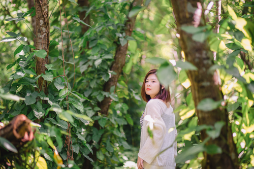 Free A woman in a white cardigan stands amidst lush green forest leaves, creating a serene natural scene. Stock Photo