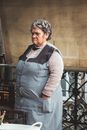 Elderly Woman Standing in an Apron and Frowning 
