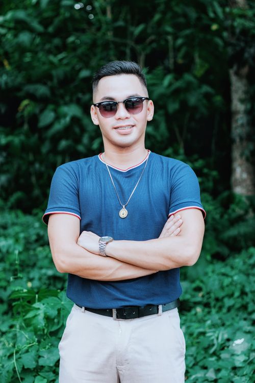 Free Smiling man in blue t-shirt with arms crossed, wearing sunglasses and necklace, set against a green plant background outdoors. Stock Photo