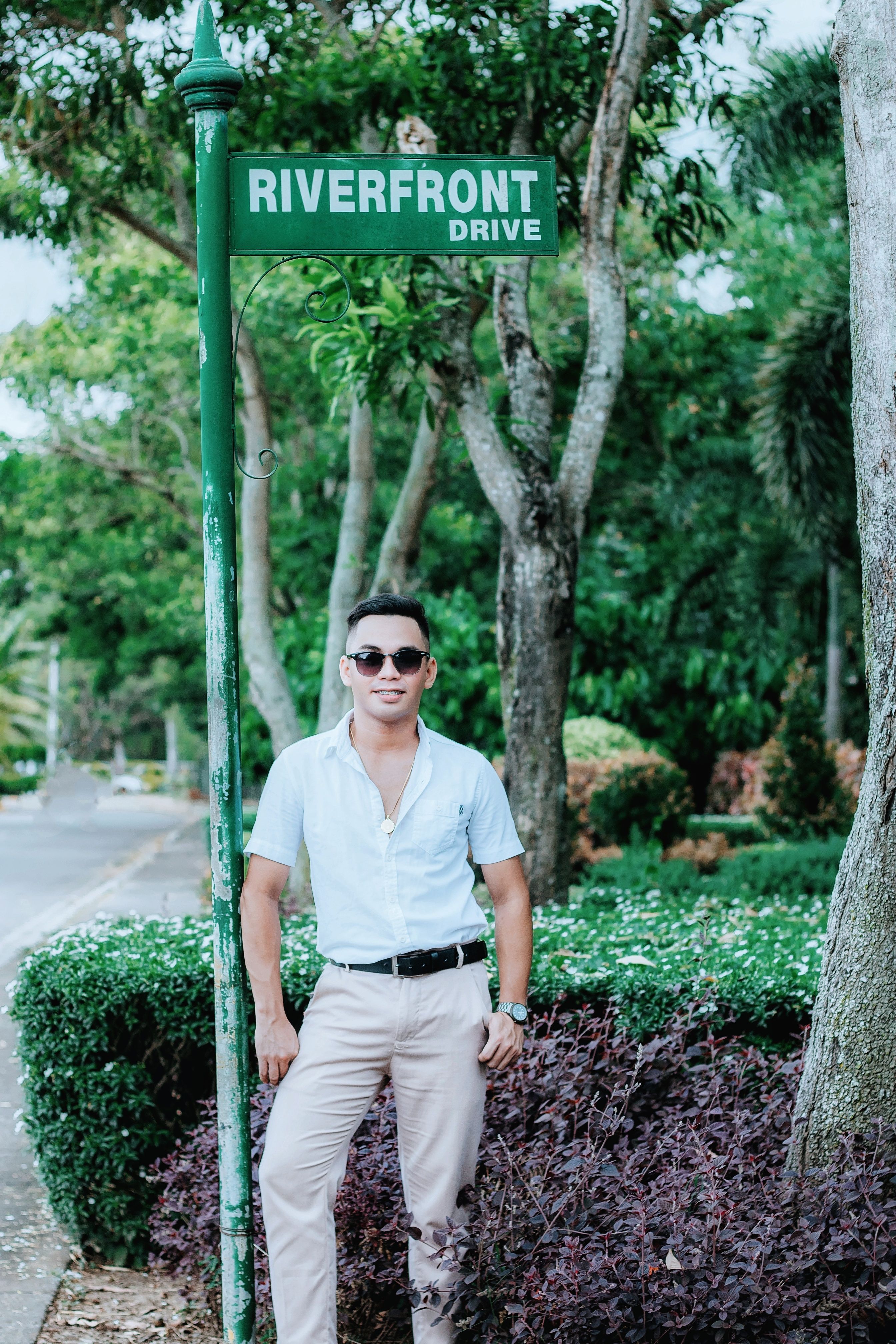 Man in sunglasses poses stylishly by a Riverfront Drive sign amidst greenery.