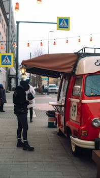 Casual city scene with a retro red food truck and customers on the sidewalk under decorative lights.
