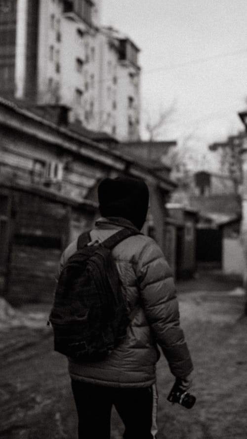 Grayscale Photo of Person in Winter Jacket Walking and Holding a Camera