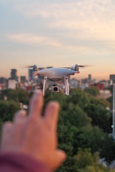 Aerial view of a white drone hovering over a cityscape with a hand reaching towards it at dusk.