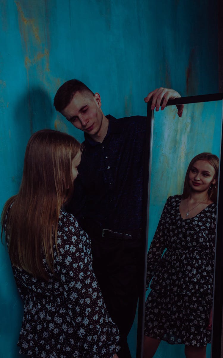 A Woman In Floral Dress Looking At Herself In The Mirror Holding By The Man In Black Long Sleeves