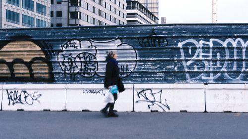 Free Person walking along graffiti-covered wall in urban street setting, heading past modern buildings. Stock Photo