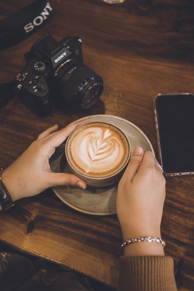 Woman Holding A Cup Of Cafe Latte And A Camera And Smart Phone On The Table 