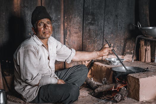 Elderly man cooking on a makeshift stove in a traditional Nepalese hut.