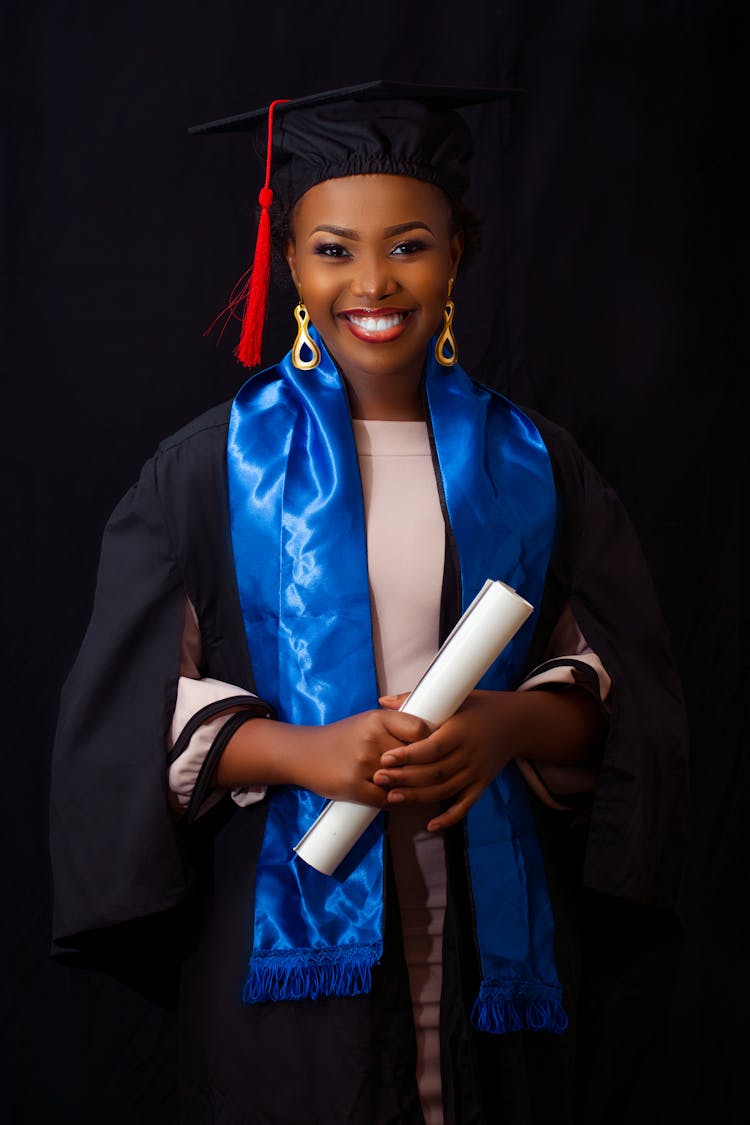 A Woman Wearing Black Graduation Gown Holding A Rolled Certificate While Smiling At The Camera