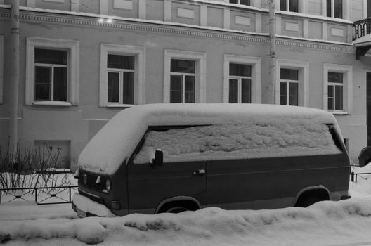 Black and white photo of a snow-covered van parked by a building in winter.