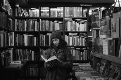 A grayscale Photo of a Woman Reading Book Inside the Library