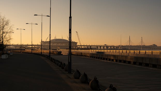 Urban landscape at sunrise featuring a bridge, stadium, and cityscape silhouette over a calm waterfront.